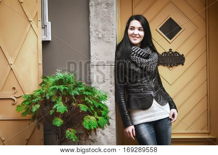 woman in black leather jacket, knit scarf and jeans standing near yellow carved wooden door with inscription welcome and smiles. Near the girls grows green fern.