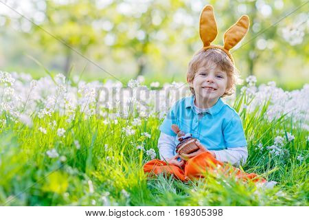 Cute happy little boy wearing Easter bunny ears at spring green grass and blooming apple garden, eating chocolate muffing with bunny and having fun outdoors.