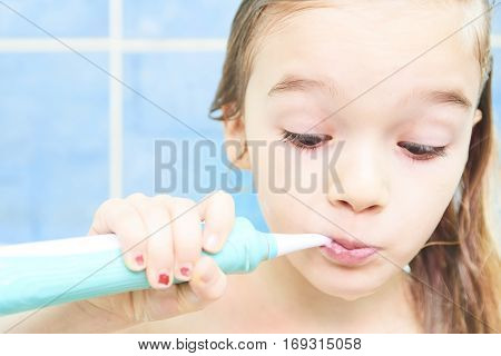 Close up of cute little girl in a bath and cleaning her teeth with electrical toothbrush against cool blue wall. Children hygiene.