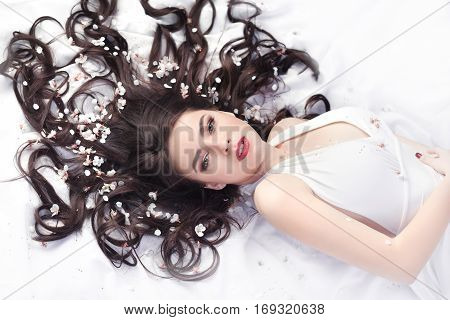 Spring beauty consept. Portrait of a fashionable model with curly brown hair sexy red lips lying on white petals background. Studio portrait shot of gorgeous woman in white dress with perfect make-up sexy posing, looking at camera.