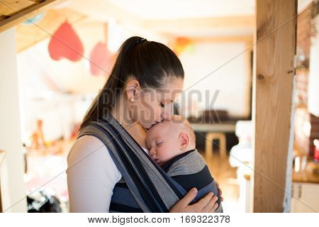 Beautiful young mother at home with her baby son sleeping in sling, kissing him on head