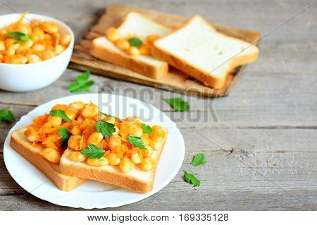Baked beans with vegetables on white bread and on plate. Baked white beans in a bowl, bread slices, parsley sprigs on wooden background with copy space for text. Healthy diet food
