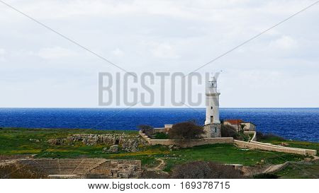 An old white lighthouse stands over the blue sea and ancient theatre ruins, in the middle of meadow coverd by grass and yelloe flowers, shooted in autumn morning