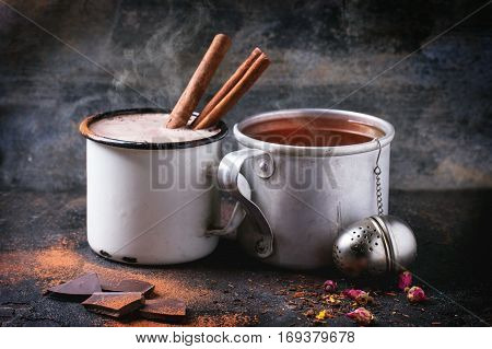 Two vintage mugs with tea and hot chocolate, served with dry tea, rose buds, chopped chocolate and cinnamon sticks over black background