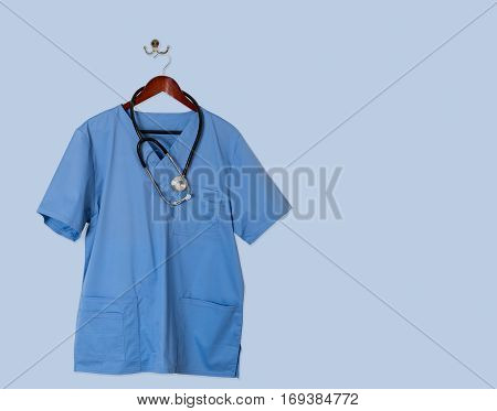 Blue medical scrubs uniform shirt hanging on a hanger with stethoscope and on hanger and hook on blue wall ready for work