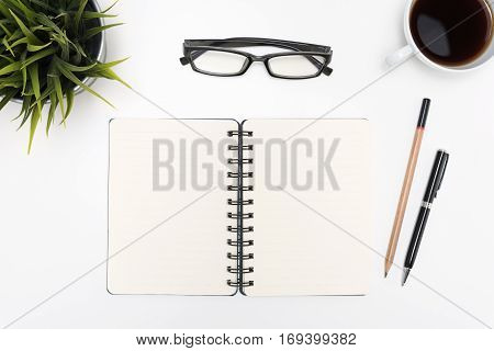 Open spiral blank notebook with pen pencil eye glasses cup of coffee and small flower plant on white desk table background flat lay top view