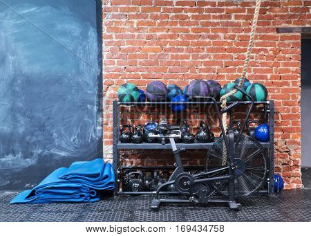 Cross-fit gym with equipment for workouts - velosimulator air bike mats and rack with kettlebells and medical balls against a red brick wall