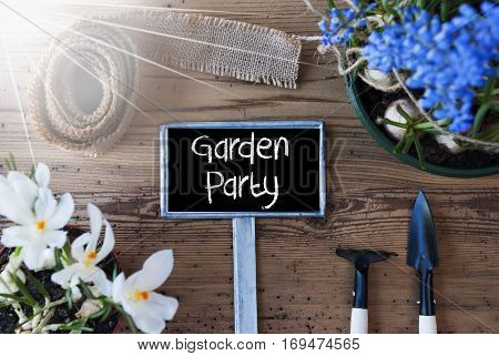Sign With English Text Garden Party. Sunny Spring Flowers Like Grape Hyacinth And Crocus. Gardening Tools Like Rake And Shovel. Hemp Fabric Ribbon. Aged Wooden Background
