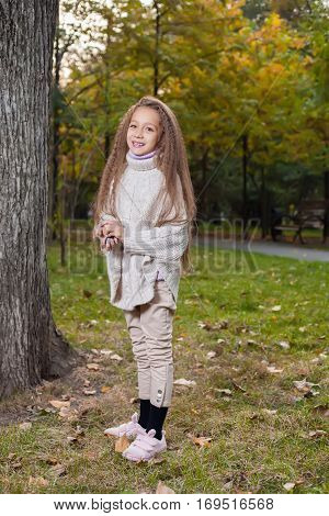 Beautiful little girl in white sweater posing in autumn Park near the tree