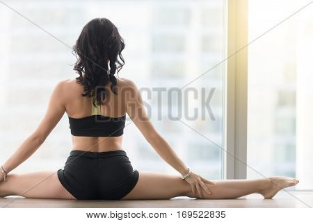 Young sporty woman practicing yoga, sitting in Samakonasana exercise, Straight Angle pose, working out, wearing sportswear, black tank top, shorts, indoor, floor window with city, rear view, closeup