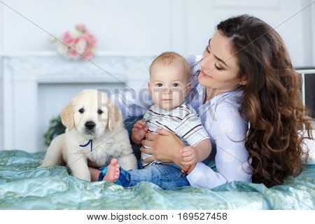 Little boy with short blond hair, barefoot, sitting on the bed on a turquoise blanket with her mother, a beautiful woman with long curly hair, black hair, wearing a white shirt and blue jeans, along with them - a pet, puppy golden retriever, happy family
