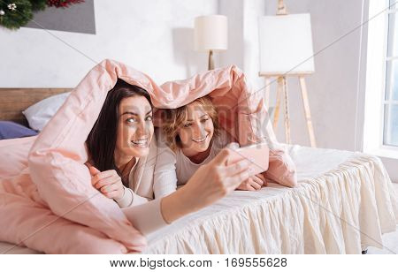 Just one shot. Joyful pretty brunette lady making a picture of herself and her charming friend while they both hiding under a pink blanket while spending Sunday morning together