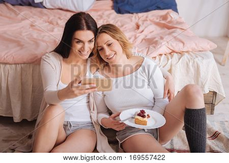 Upload it somewhere. Two charming happy beautiful ladies taking a selfie using a smartphone to show their friends their pleasant moments together