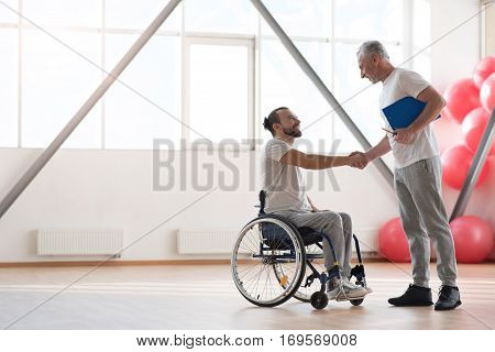Nice to meet you. Happy positive smiling disabled man sitting in the wheelchair while shaking hands with his trainer