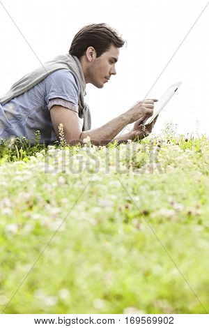 Side view of young man using digital tablet while lying on grass against clear sky