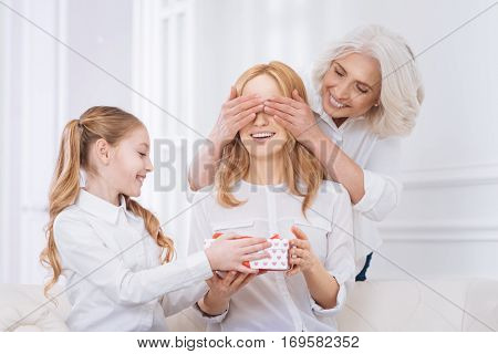 What a surprise. Cheerful content adult woman sitting on the couch while her mother and daughter congratulating her while going to celebrate at home