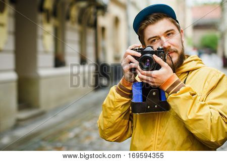Young hipster with a beard holding oldshool film camera, thinking and ready to take a photo, on the street, wearing in bright raincoat and hat, portrait, day outdoors, closeup, soft-focus, sly glance