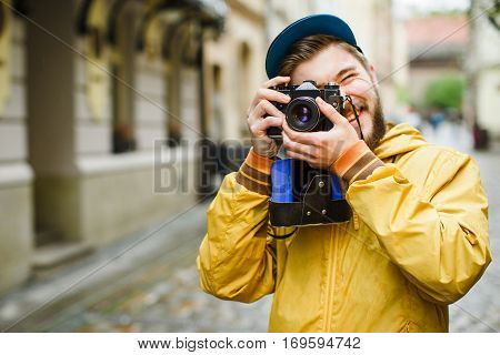 Young hipster with a beard holding oldshool film camera, looking in the camera viewfinder, laughing and ready to take a photo, on the street, wearing in bright raincoat and hat, portrait, day outdoors, closeup