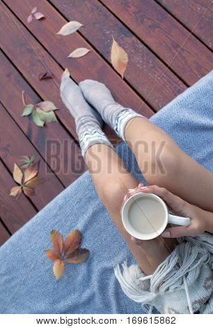 Woman with cup of coffee sitting relaxing on patio wooden deck, she's covered in a blanket. Fall leaves on wooden patio deck. Autumn concept.