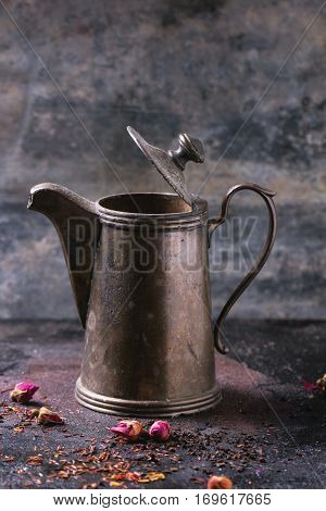 Open vintage teapot with dry tea and rose buds over black metal background