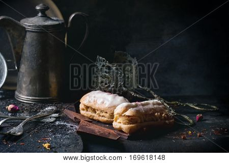 Tea drinking with eclairs and chopped chocolate, served with vintage teapot and cutlery over dark background. See series