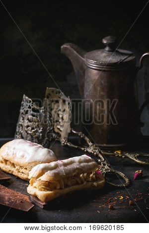 Tea drinking with eclairs and chopped chocolate, served with vintage teapot and cutlery over dark background. See series