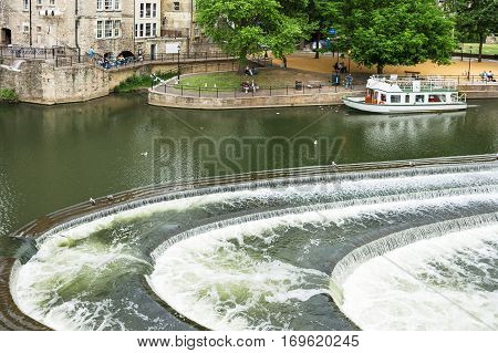 Bath Somerset United Kingdom - June 19 2006: People relaxing in the park near the Pulteney Bridge. The river Avon with weir and tour boat in Bath.