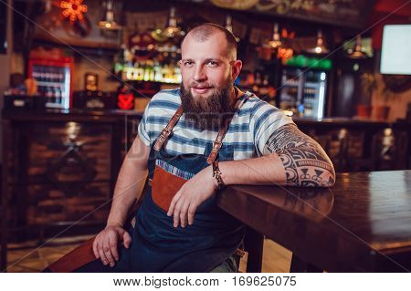 Bearded barman with tattoos and wathes wearing an apron sitting in bar.