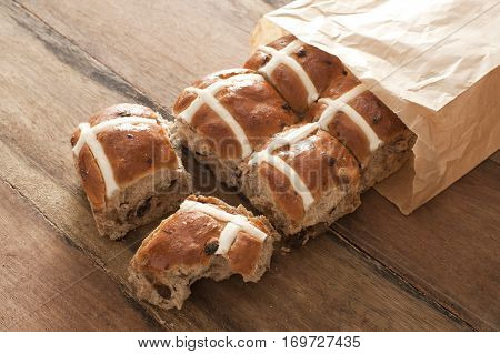 Hot Cross Buns for Easter in a brown paper packet spilling out onto a wooden table with one bun having a missing bite