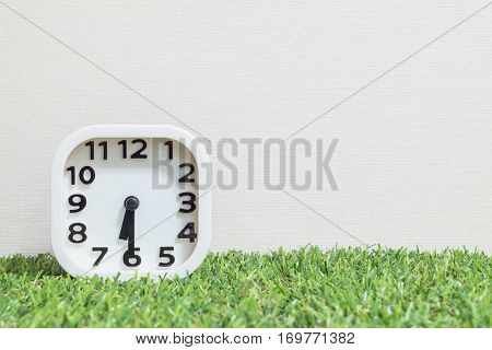 Closeup white clock for decorate show a half past six or 6:30 a.m. on green artificial grass floor and cream wallpaper textured background with copy space