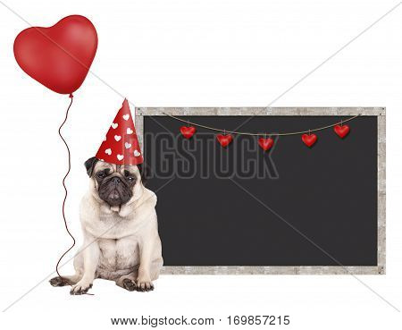 cute pug puppy dog with red party hat sitting next to blank blackboard sign and holding heart shaped balloon isolated on white background