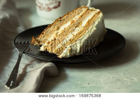 Slice of homemade carrot and honey cake with cream cheese frosting on a dark plate, cup of coffe or tea, cozy atmosphere
