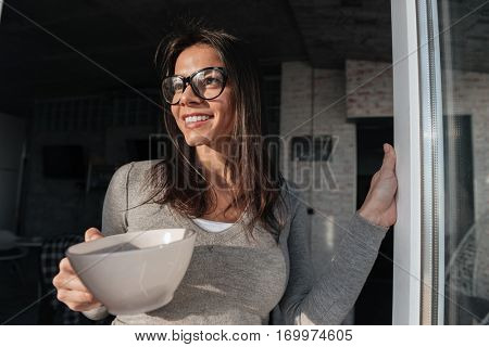 Picture of young cheerful woman wearing eyeglasses drinking coffee indoors. Look aside.