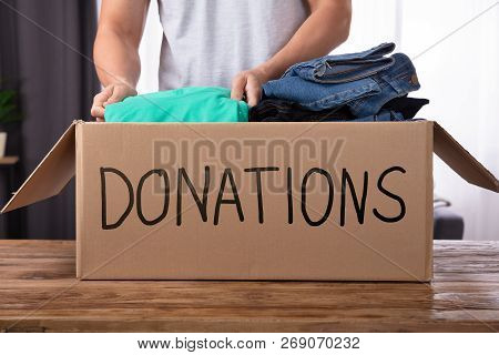Young Man Donating Clothes In Donation Box Over Wooden Desk