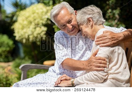Senior man in medical patient cloth hugging wife outdoor bench. Old woman is consoled by her husband suffering from a terminal illness. Wife visit her husband at the hospital after the operation.