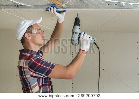 Young Man In Goggles Fixing Drywall Suspended Ceiling To Metal Frame Using Electrical Screwdriver On