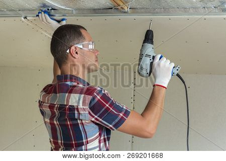 Young Man In Goggles Fixing Drywall Suspended Ceiling To Metal Frame Using Electrical Screwdriver On