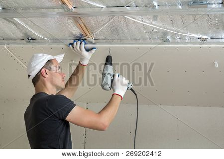 Young Man In Goggles Fixing Drywall Suspended Ceiling To Metal Frame Using Electrical Screwdriver On