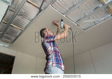 Young Man In Usual Clothing And Work Gloves Fixing Drywall Suspended Ceiling To Metal Frame Using El