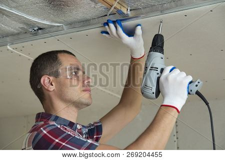 Young Man In Goggles Fixing Drywall Suspended Ceiling To Metal Frame Using Electrical Screwdriver On