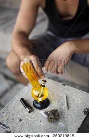 Young Man Smoking Pot Using Bong; Detail Of Male Hand Filling Up Bong With Cannabis, Getting It Read