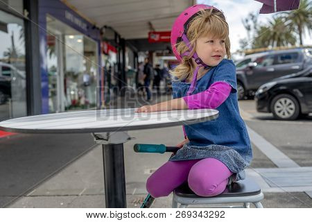 Young girl in pink outfit and pink helmet riding a kick-scooter sitting at the outside cafe table wi