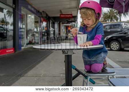 Young Girl In Pink Outfit And Pink Helmet Riding A Kick-scooter Sitting At The Outside Cafe Table Wi
