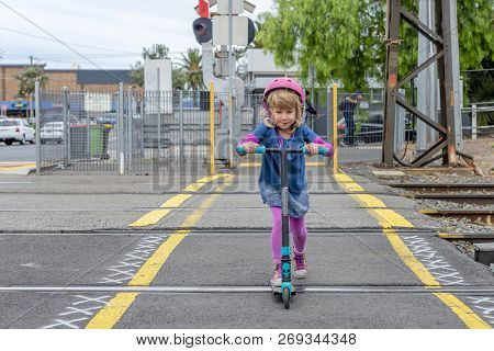 Young Girl In Pink Outfit And Pink Helmet Riding A Kick-scooter Across The Railway Line