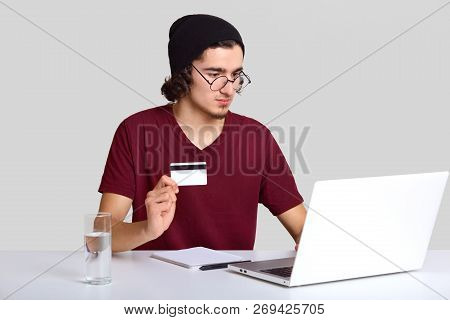 Studio Shot Of Serious Guy Looks Attentively At Monitor Of Laptop, Holds Plastic Card, Makes Money T