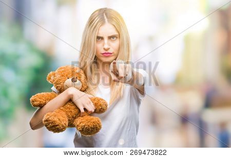 Young beautiful blonde woman holding teddy bear over isolated background pointing with finger to the camera and to you, hand sign, positive and confident gesture from the front