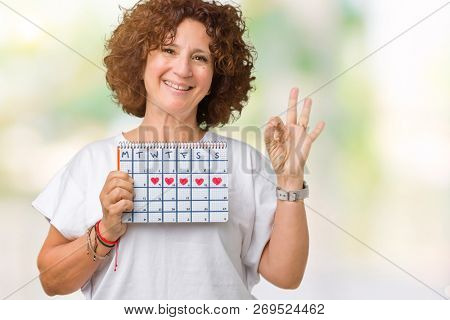 Middle ager senior woman holding menstruation calendar over isolated background doing ok sign with fingers, excellent symbol