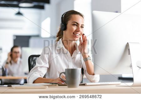 Image of a happy emotional business woman in office callcenter working with computer wearing headphones.