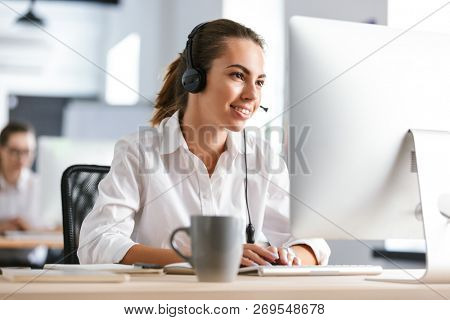 Photo of a happy cheerful business woman in office callcenter working with computer wearing headphones.