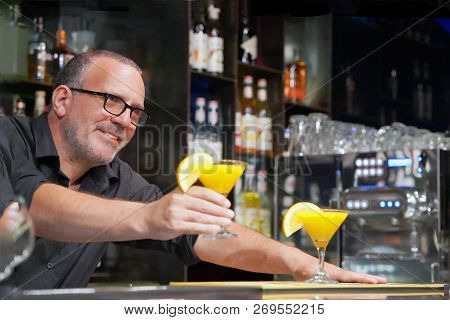 The Barman Gives A Cocktail To The Client Of The Hotel Bar. Fresh Yellow Cocktail With Orange. Alcoh